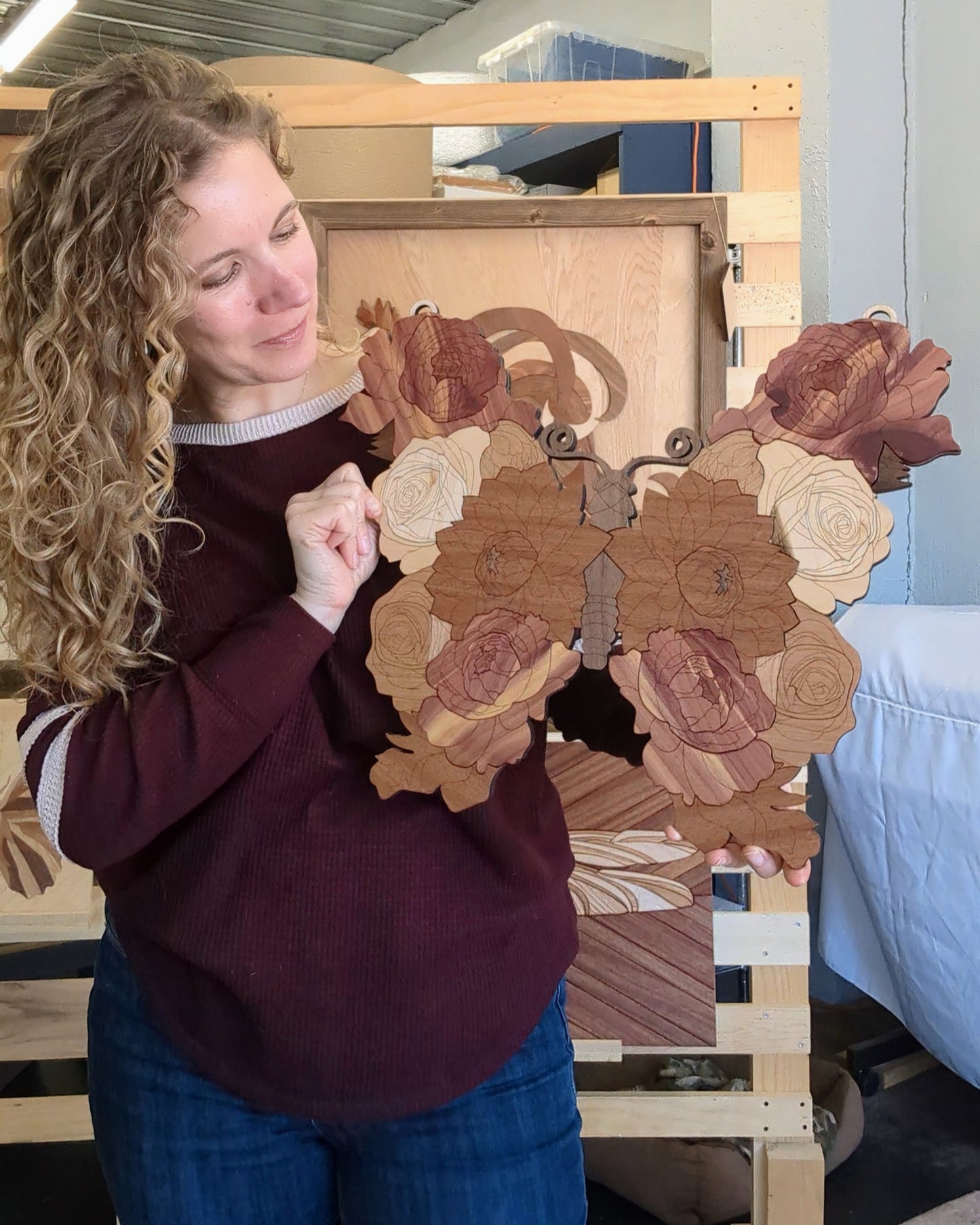 Woman holding a handcrafted wooden butterfly, looking down at it and smiling softly. The piece features layered floral details and intricate cuts.