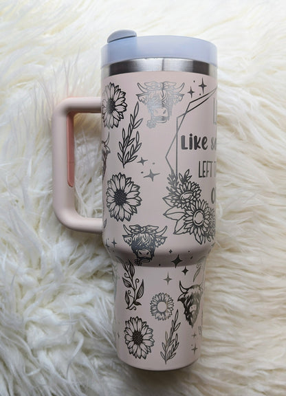 Photo of a soft pink colored 40 oz insulated tumbler with handle permanently engraved with a highland cow pattern and the text Live Like Someone Left the Gate Open