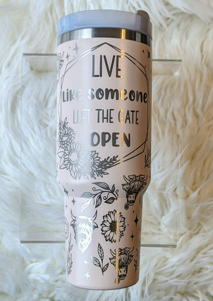 Photo of a soft pink colored 40 oz insulated tumbler with handle permanently engraved with a highland cow pattern and the text Live Like Someone Left the Gate Open
