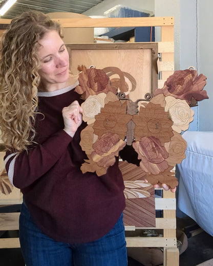 Woman holding a handcrafted wooden butterfly, looking down at it and smiling softly. The piece features layered floral details and intricate cuts.