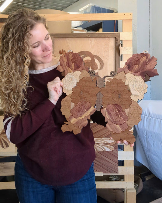 Woman holding a handcrafted wooden butterfly, looking down at it and smiling softly. The piece features layered floral details and intricate cuts.