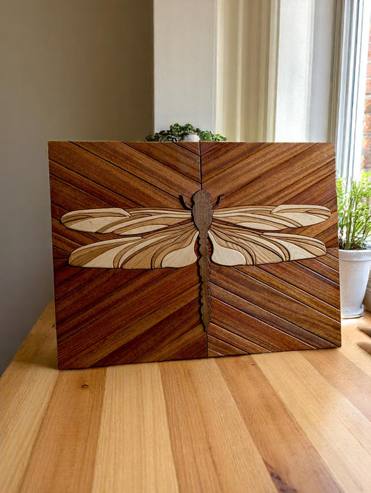 Hardwood dragonfly wall art staged in natural light on a wood surface with a potted plant in the background.